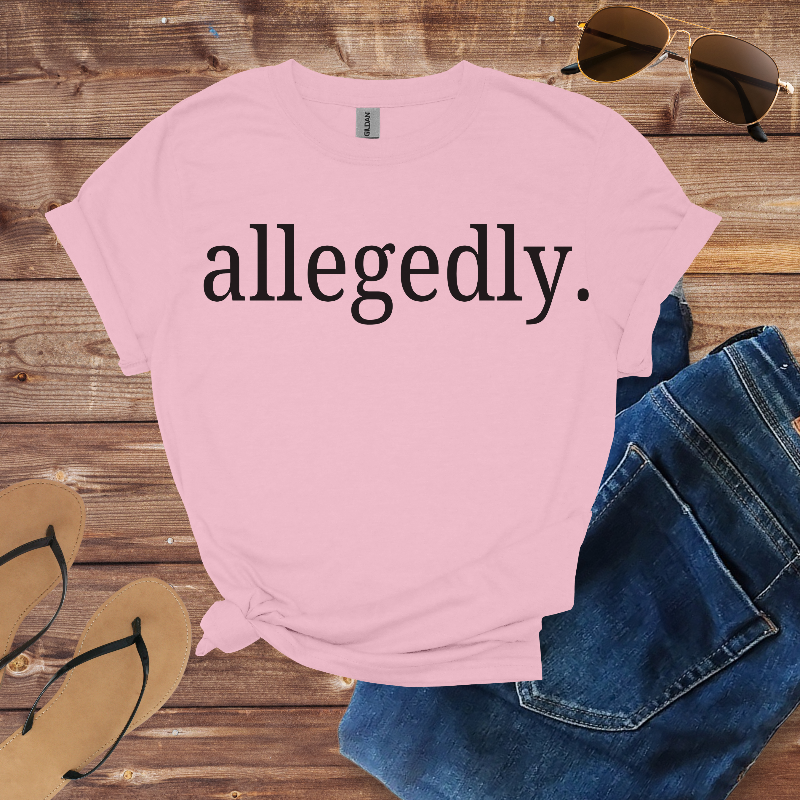 Pink t-shirt with 'allegedly.' text on a wooden surface with sunglasses and jeans.