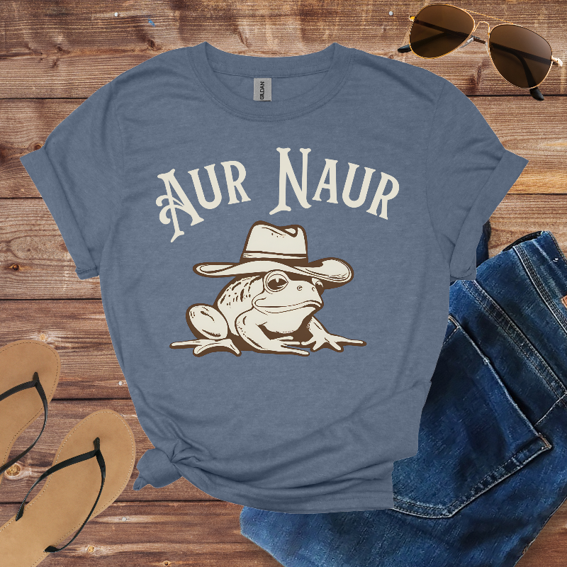 Blue t-shirt with a frog wearing a hat graphic and 'Aur Naur' text, placed on a wooden surface with sunglasses, jeans, and sandals.
