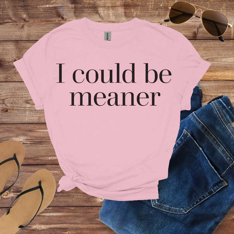 Pink t-shirt with 'I could be meaner' text on a wooden surface with sunglasses and jeans.