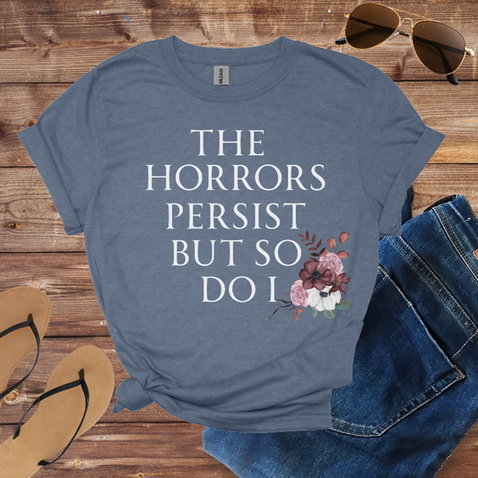 Blue t-shirt with 'The Horrors Persist But So Do I' text and floral design on a wooden surface with jeans and sandals.