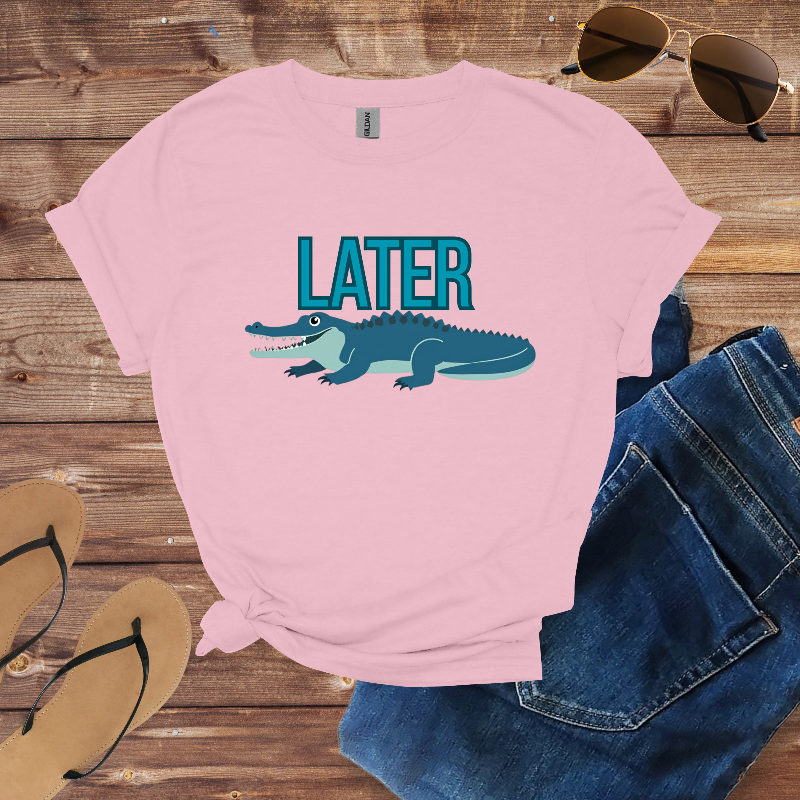 Pink t-shirt with alligator graphic and 'LATER' text on a wooden surface with sunglasses and flip-flops.