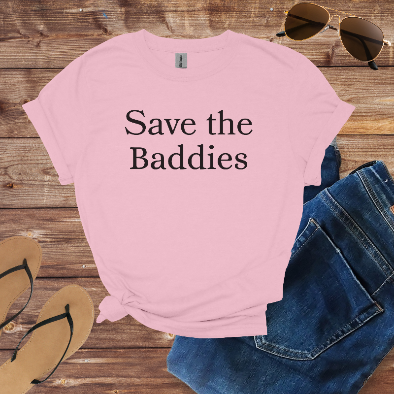 Pink t-shirt with 'Save the Baddies' text on a wooden surface with sunglasses and jeans.