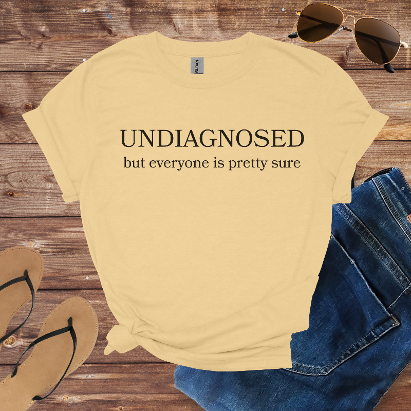 Beige t-shirt with 'undiagnosed but everyone is pretty sure' text on a wooden surface with sunglasses, jeans, and flip-flops.