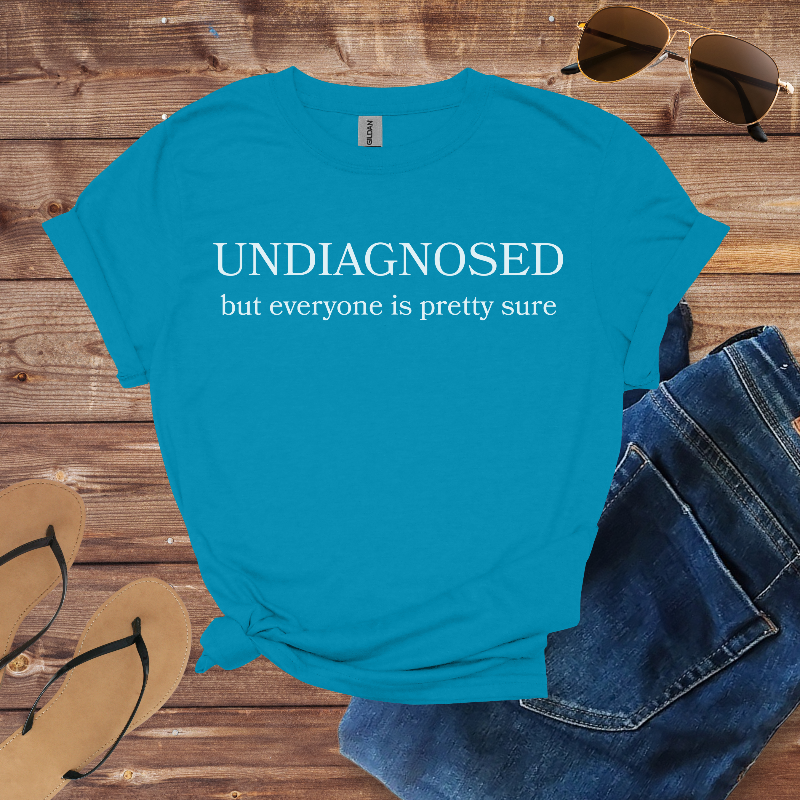 Blue t-shirt with 'undiagnosed but everyone is pretty sure' text on a wooden surface with sunglasses and jeans.