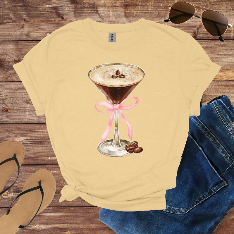 Beige t-shirt with a coffee-themed design on a wooden surface with sunglasses, jeans, and sandals.
