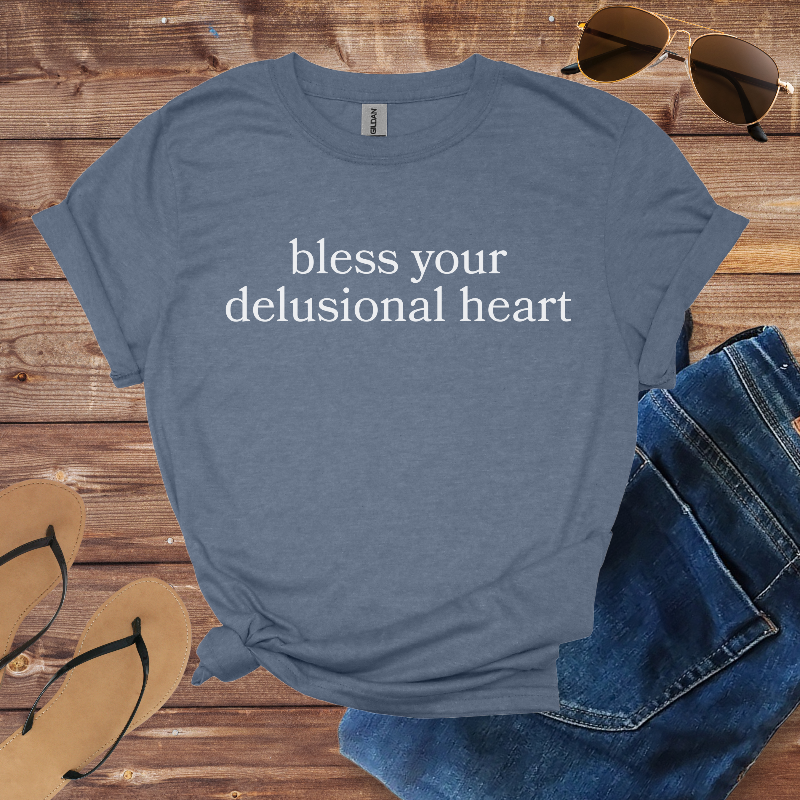 Blue t-shirt with 'bless your delusional heart' text on a wooden surface with sunglasses, jeans, and sandals.