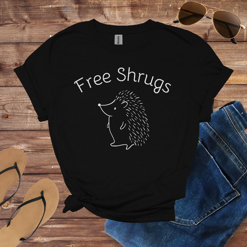 Black t-shirt with 'Free Shrugs' and hedgehog graphic on a wooden surface with sunglasses, jeans, and sandals.