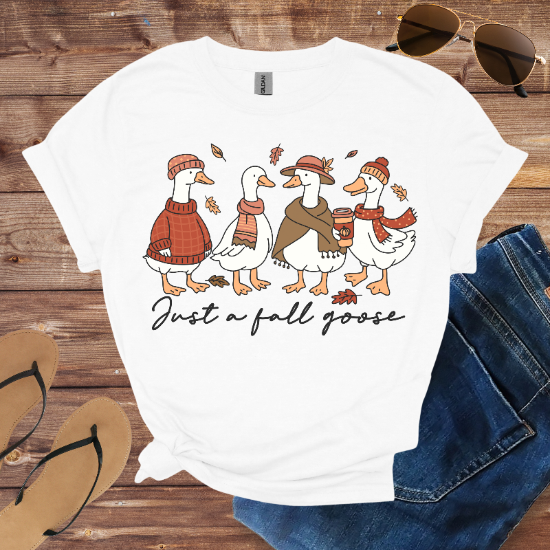 White t-shirt with cartoon ducks wearing fall clothing and 'Just a fall goose' text on a wooden surface with jeans and sunglasses.