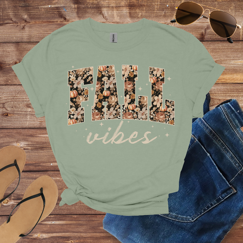 Green t-shirt with 'FALL vibes' design on a wooden surface with sunglasses, jeans, and sandals.