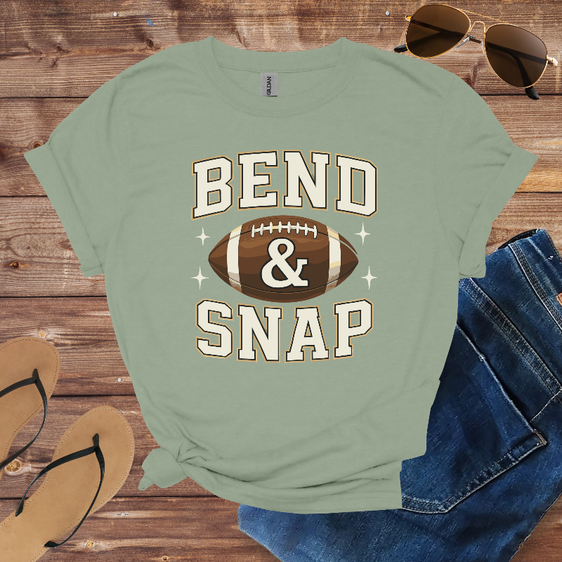 Green t-shirt with 'Bend & Snap' and football graphic on a wooden surface with sunglasses, jeans, and sandals.
