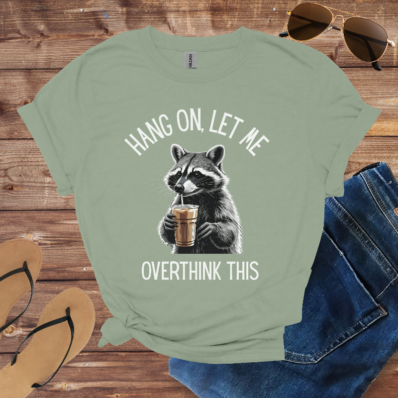 Green t-shirt with raccoon graphic and text on a wooden surface with sunglasses, flip-flops, and jeans.