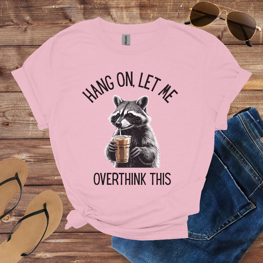 Pink t-shirt with raccoon graphic and text on a wooden surface with sunglasses and jeans.