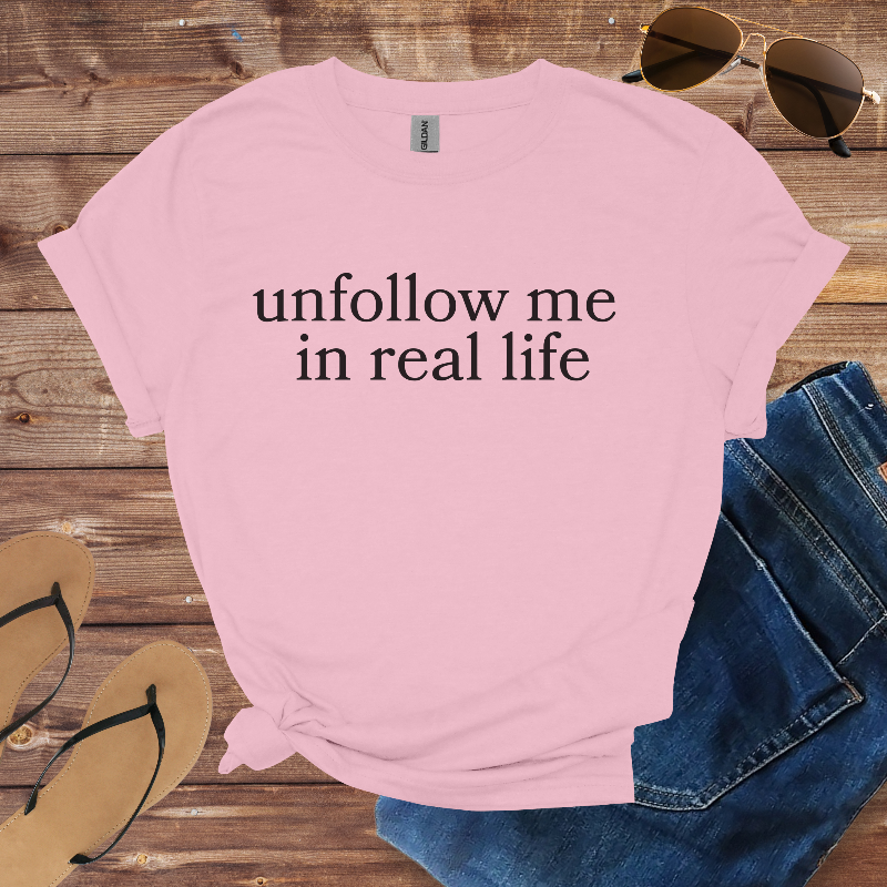 Pink t-shirt with 'unfollow me in real life' text on a wooden surface with sunglasses, jeans, and sandals.