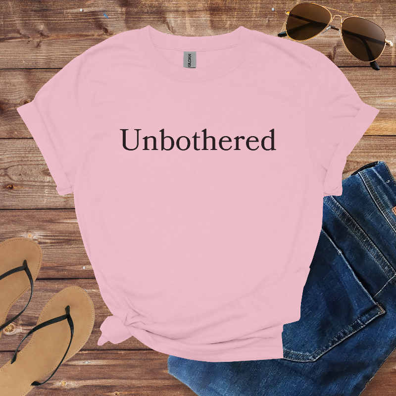 Pink t-shirt with 'Unbothered' text on a wooden surface with sunglasses, jeans, and flip-flops.