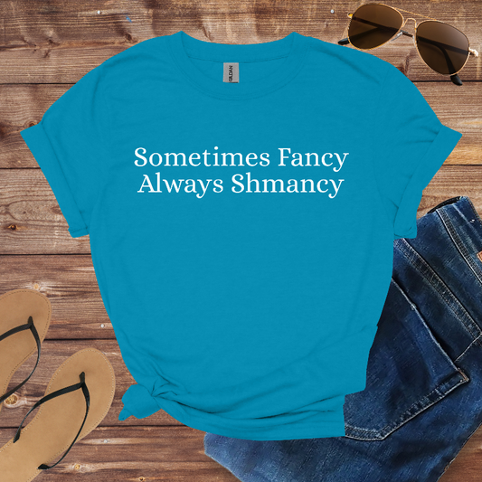 Blue t-shirt with 'Sometimes Fancy Always Shmancy' text on a wooden surface with sunglasses, jeans, and flip-flops.