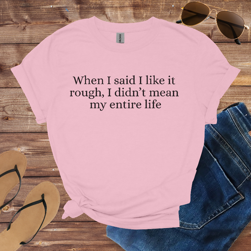 Pink t-shirt with humorous text on a wooden surface with sunglasses, jeans, and flip-flops.
