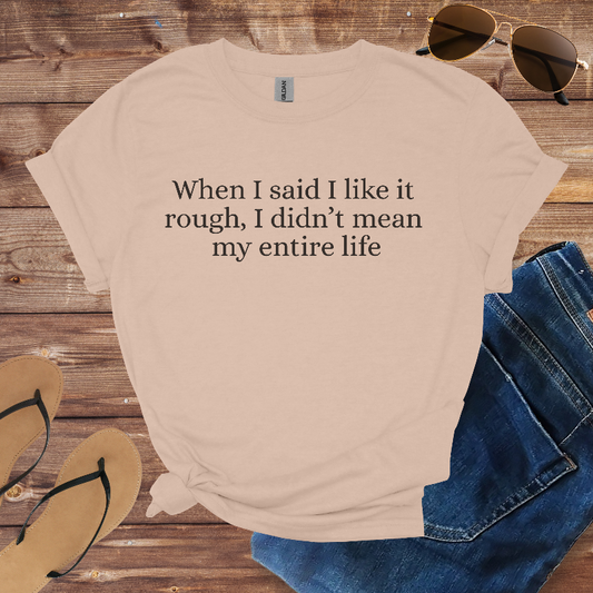 Beige t-shirt with humorous text on a wooden surface with sunglasses, jeans, and flip-flops.
