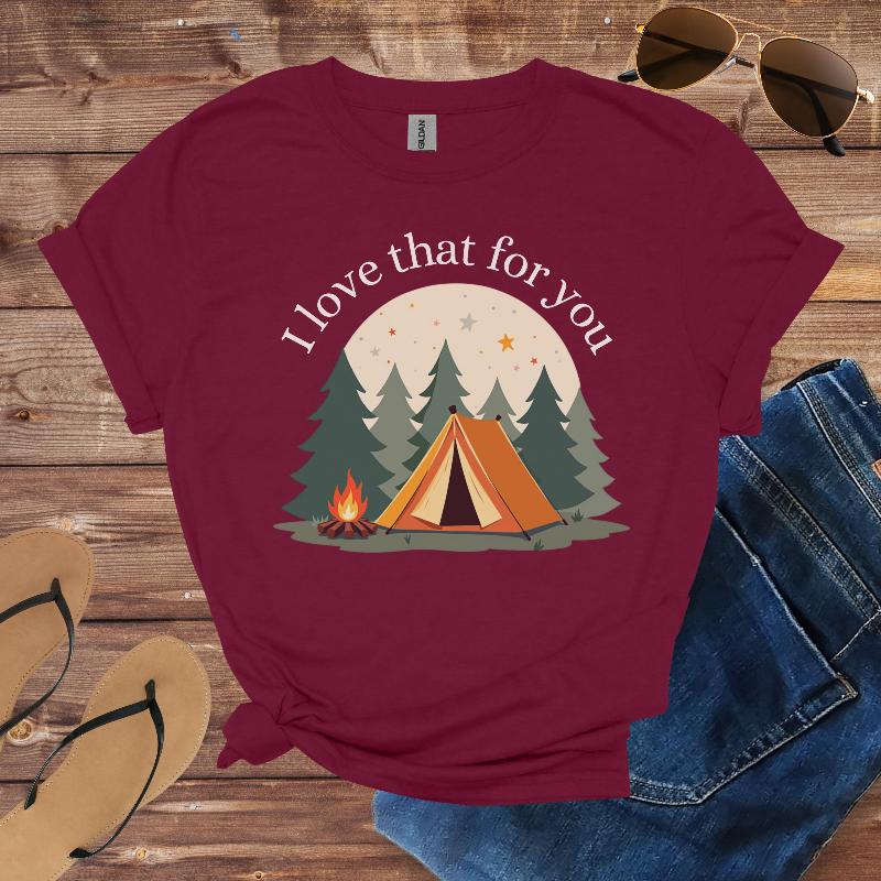 Maroon t-shirt with camping graphic and text on a wooden surface with sunglasses, flip-flops, and jeans.