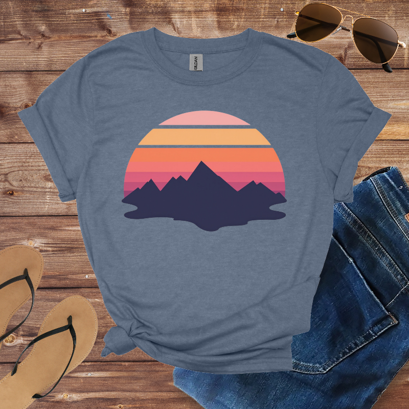 Blue t-shirt with mountain and sunset design on a wooden surface with sunglasses, jeans, and sandals.