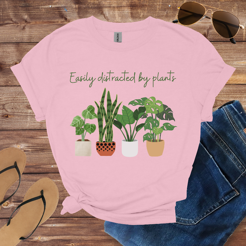 Pink t-shirt with plant illustration and text 'Easily distracted by plants' on a wooden surface with sunglasses, jeans, and sandals.