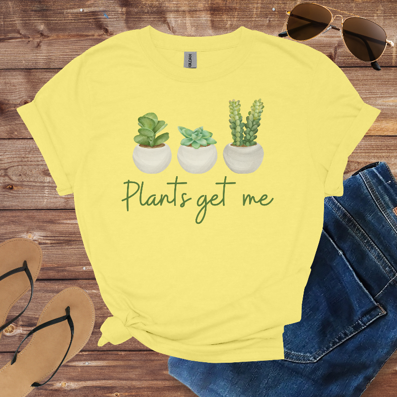 Yellow t-shirt with plant graphic and text 'Plants get me' on a wooden surface with sunglasses, jeans, and sandals.
