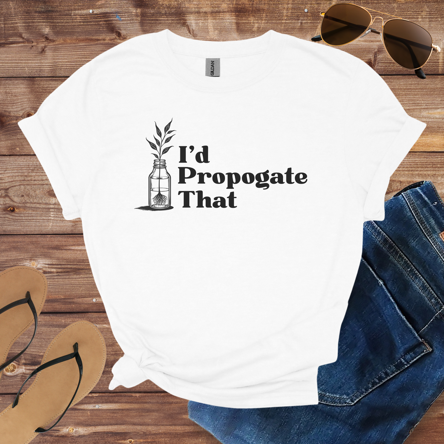 White t-shirt with 'I'd Propogate That' text and plant illustration on a wooden surface with sunglasses, jeans, and flip-flops.