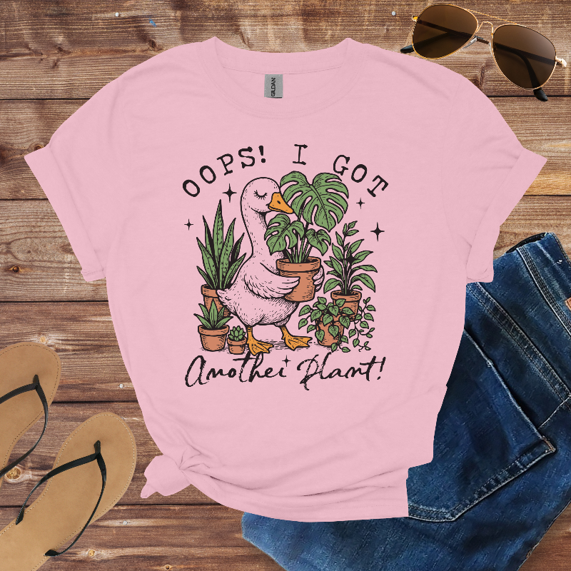 Pink t-shirt with a graphic of a duck holding plants and text 'Oops! I Got Another Plant!' on a wooden surface with sunglasses and jeans.