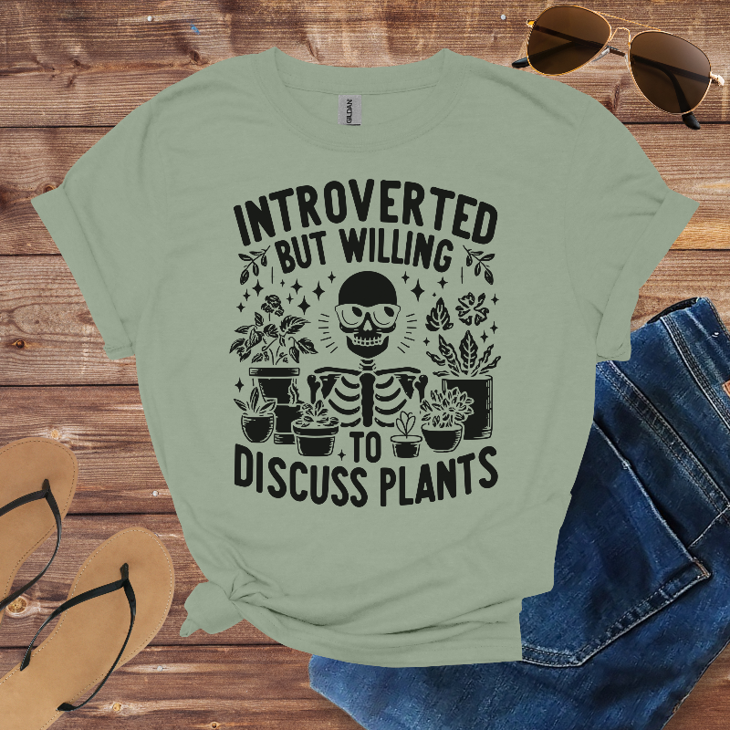 Light green t-shirt with skeleton and plants graphic on a wooden surface with sunglasses, flip-flops, and jeans.