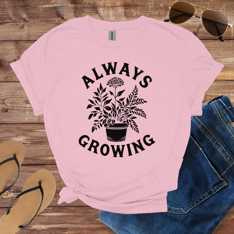 Pink t-shirt with 'Always Growing' plant graphic on a wooden surface with sunglasses and jeans.