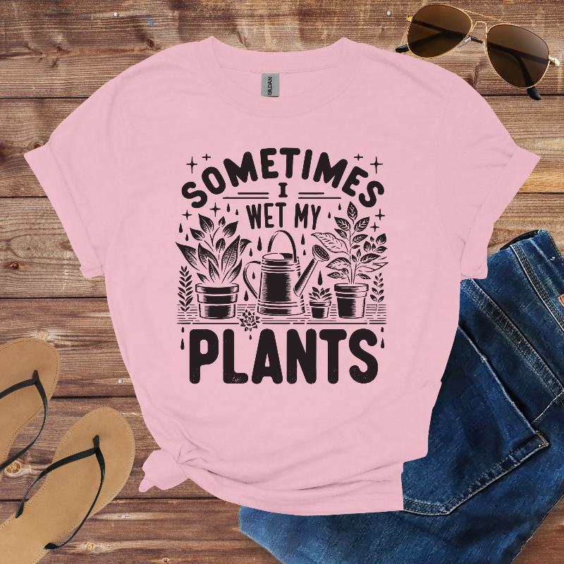 Pink t-shirt with 'Sometimes I Wet My Plants' text and graphics on a wooden surface with sunglasses, jeans, and sandals.