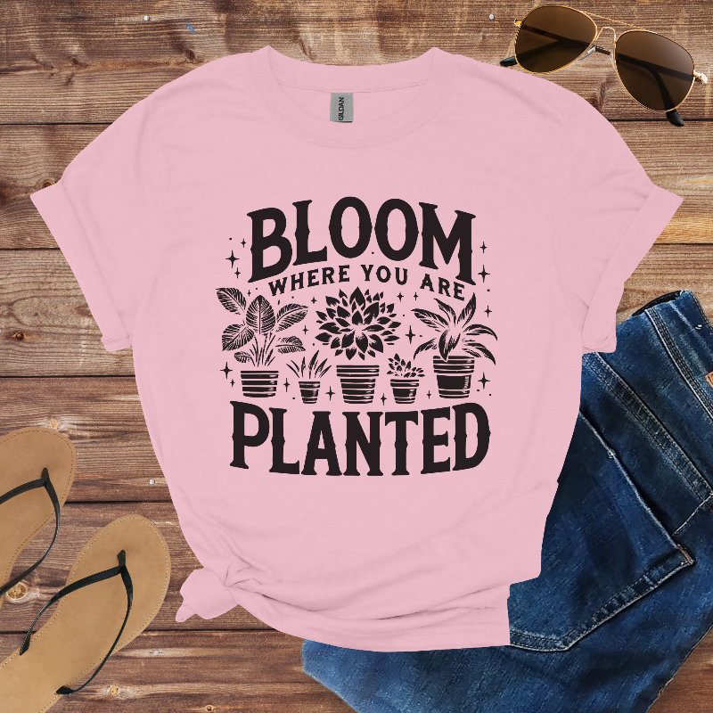 Pink t-shirt with 'Bloom where you are planted' text and plant graphics on a wooden surface with sunglasses, jeans, and sandals.