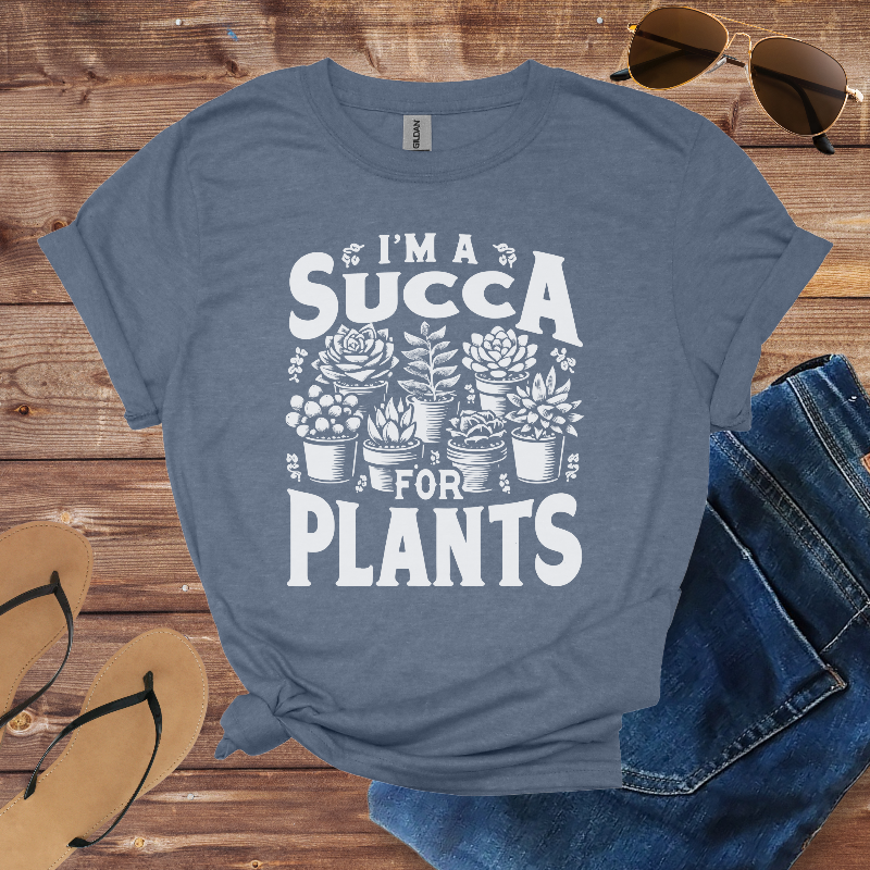 Blue t-shirt with 'I'm a Succa for Plants' design on a wooden surface with sunglasses, jeans, and sandals.