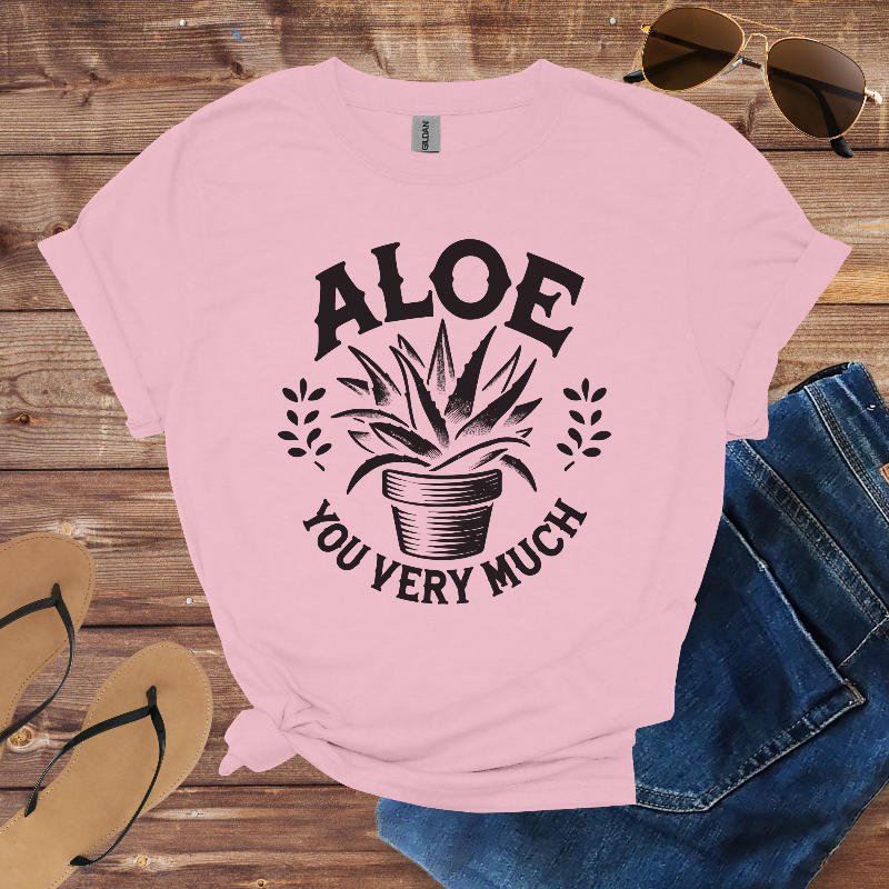 Pink t-shirt with 'Aloe You Very Much' design on a wooden surface with sunglasses, jeans, and flip-flops.