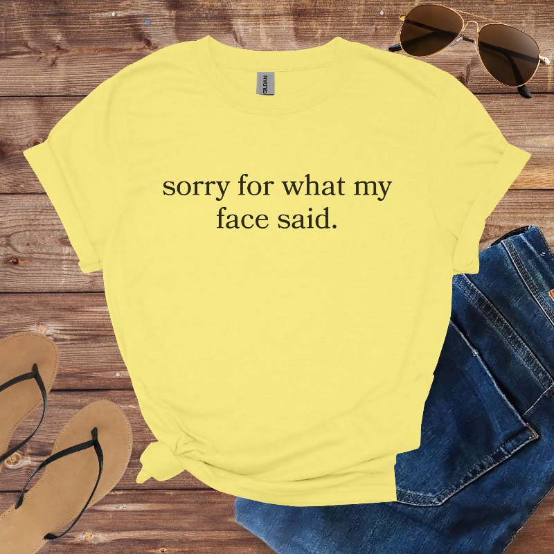Yellow t-shirt with 'sorry for what my face said' text on a wooden surface with sunglasses, jeans, and sandals.