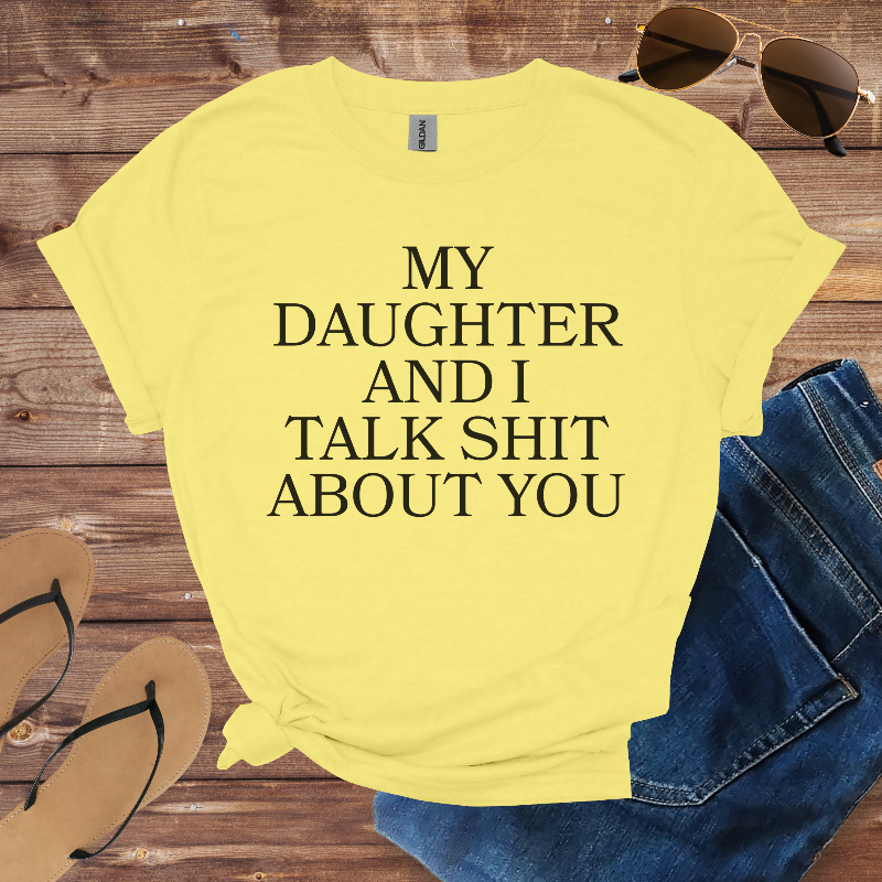 Yellow t-shirt with text 'My daughter and I talk shit about you' on a wooden surface with sunglasses and jeans.