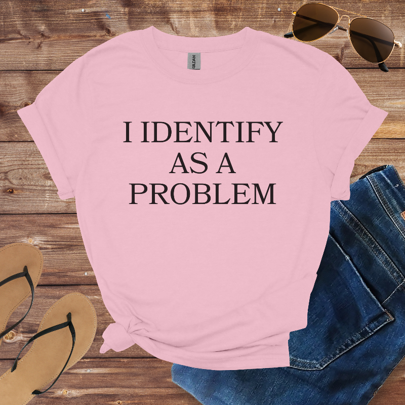 Pink t-shirt with 'I IDENTIFY AS A PROBLEM' text on a wooden surface with sunglasses, jeans, and flip-flops.