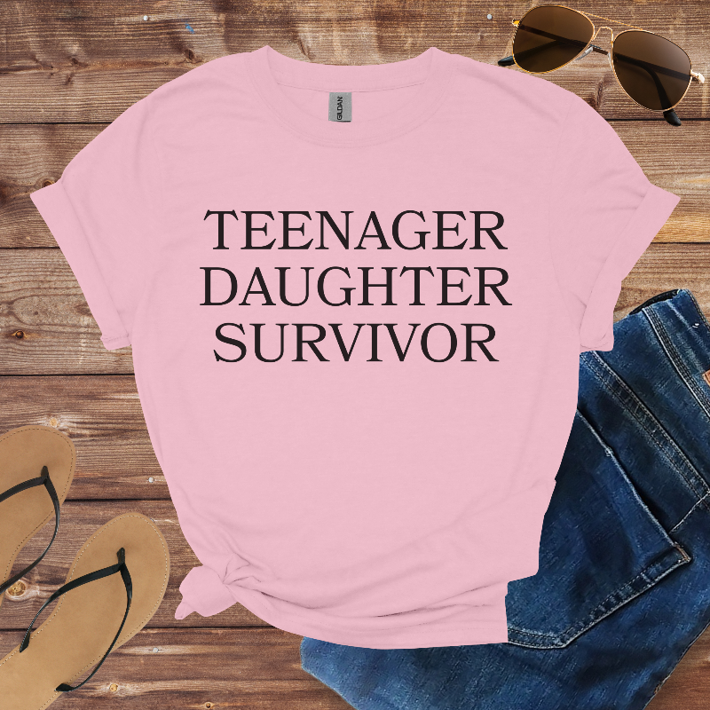 Pink t-shirt with 'TEENAGER DAUGHTER SURVIVOR' text on a wooden surface with sunglasses and jeans.