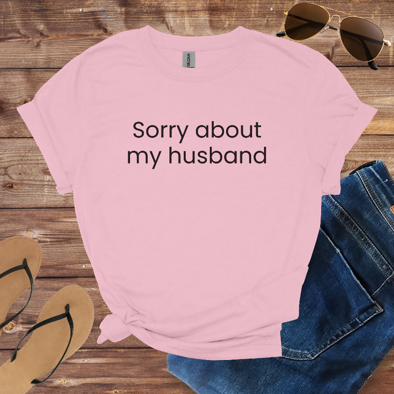Pink t-shirt with 'Sorry about my husband' text on a wooden surface with sunglasses, jeans, and flip-flops.