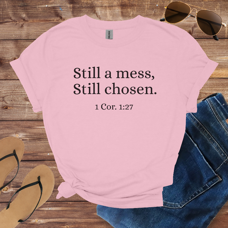 Pink t-shirt with text 'Still a mess, Still chosen. 1 Cor. 1:27' on a wooden surface with sunglasses, jeans, and sandals.