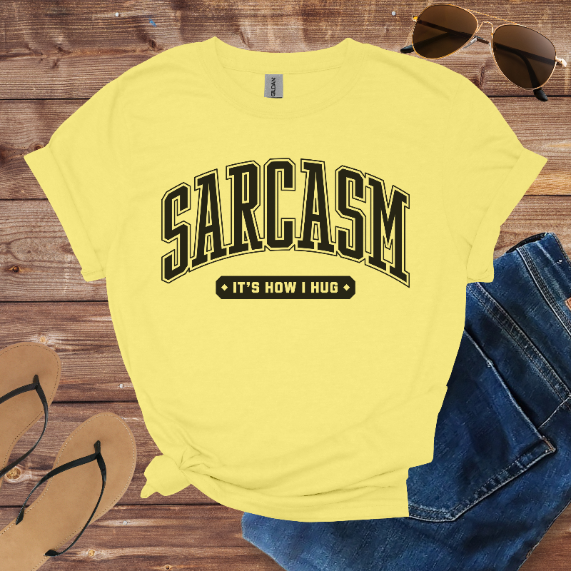 Yellow t-shirt with 'SARCASM IT'S HOW I HUG' text on a wooden surface with sunglasses, jeans, and sandals.