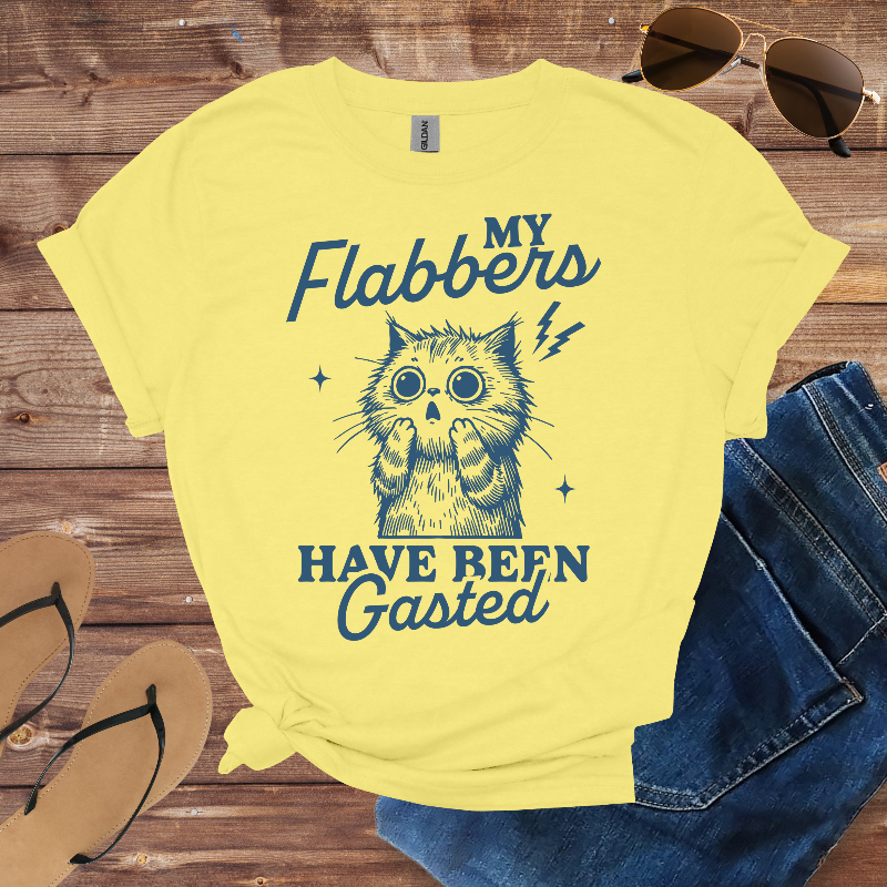 Yellow t-shirt with a cat graphic and text on a wooden surface with sunglasses and jeans.