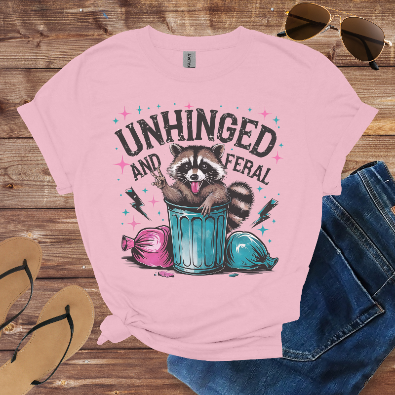 Pink t-shirt with raccoon graphic and text 'Unhinged and Feral' on a wooden surface with sunglasses and jeans.
