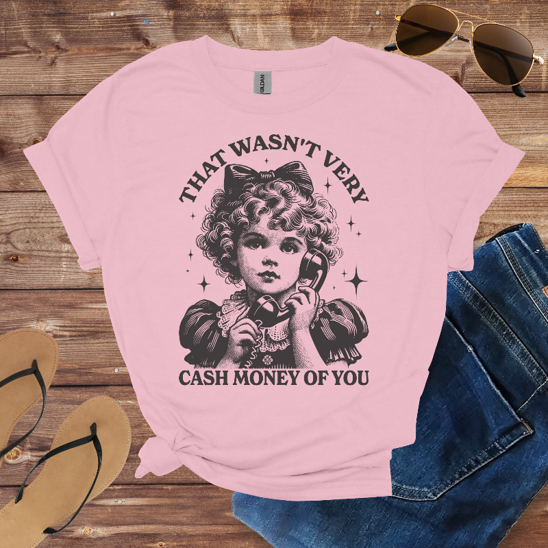 Pink t-shirt with vintage-style graphic and text on a wooden surface with sunglasses and jeans.
