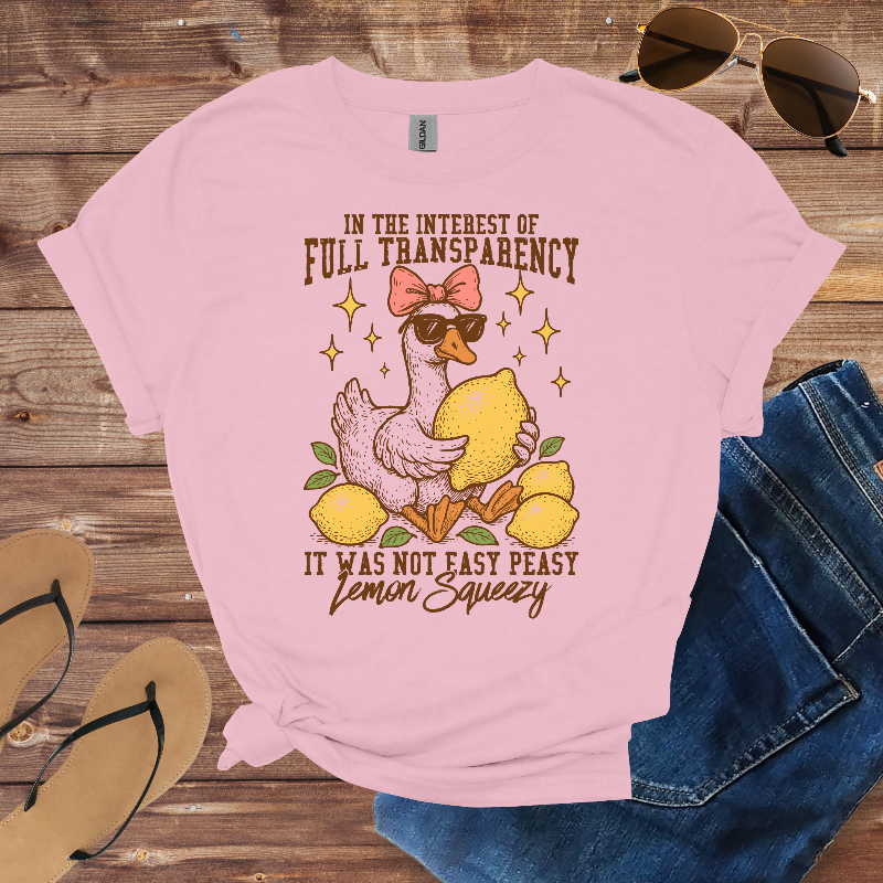 Pink t-shirt with a graphic of a duck holding lemons and text on a wooden surface with sunglasses and jeans.