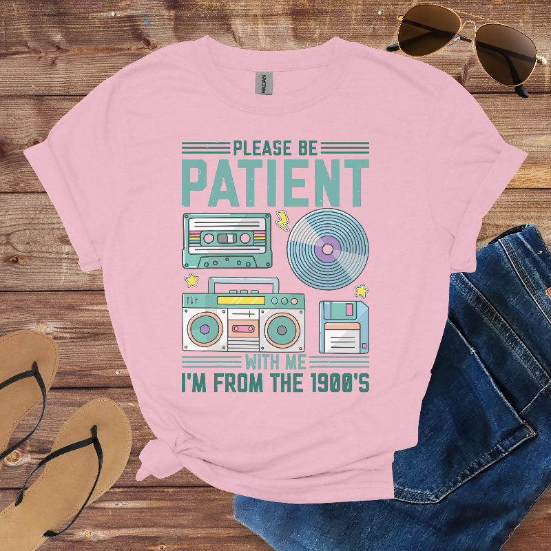 Pink t-shirt with retro design and text on a wooden surface with sunglasses, jeans, and flip-flops.