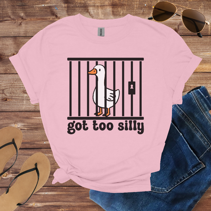 Pink t-shirt with a goose graphic and text 'got too silly' on a wooden surface with sunglasses, jeans, and sandals.
