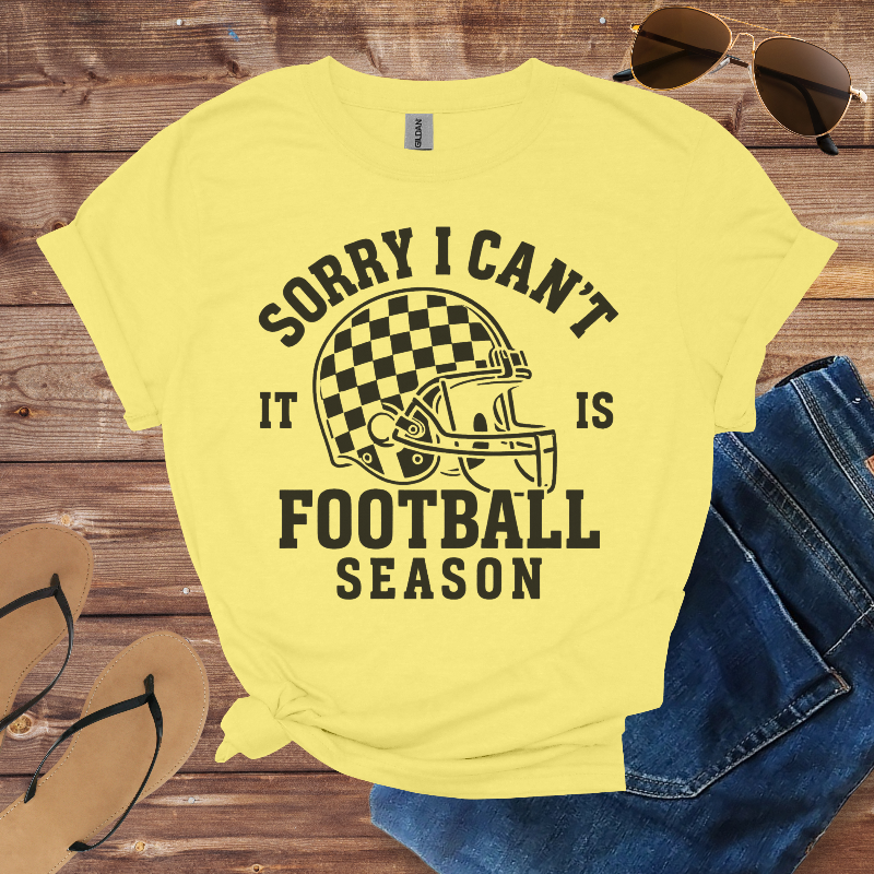 Yellow t-shirt with football-themed text and graphics on a wooden surface with sunglasses and jeans.