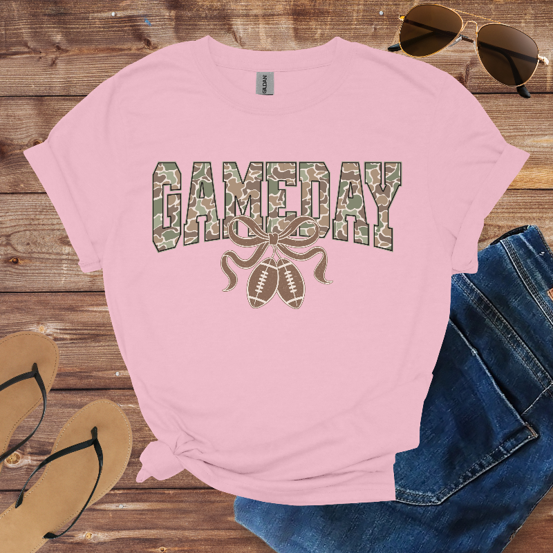 Pink t-shirt with 'GAMEDAY' print on a wooden surface with sunglasses, jeans, and sandals.