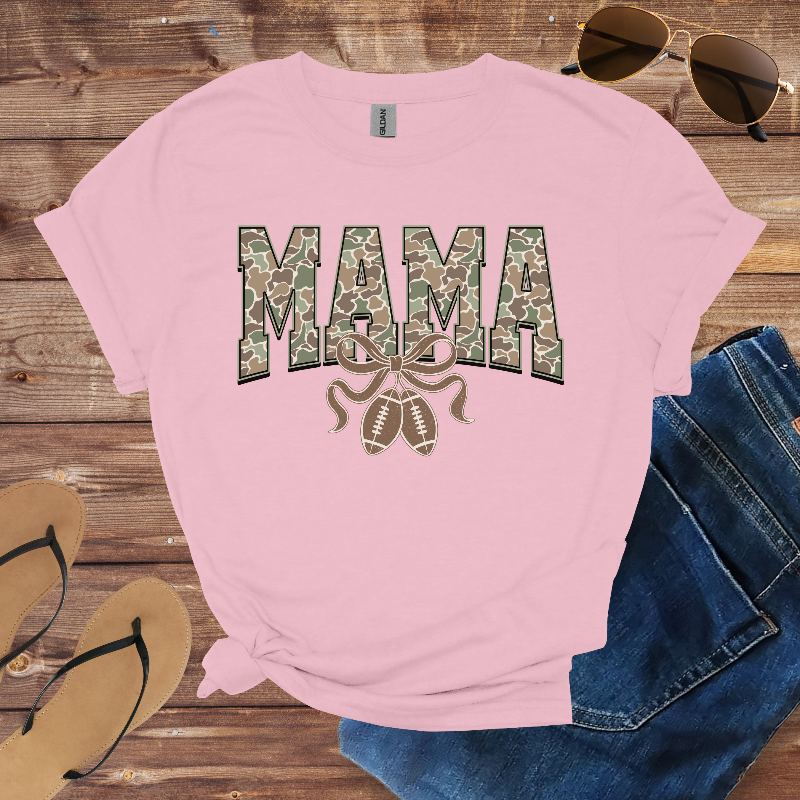Pink t-shirt with 'MAMA' and camouflage design on a wooden surface with sunglasses, jeans, and sandals.