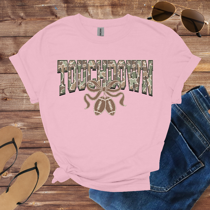 Pink t-shirt with 'TOUCHDOWN' graphic on a wooden surface with sunglasses, jeans, and flip-flops.